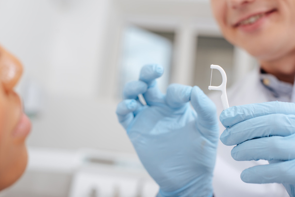 Ausgeschnittene Ansicht eines Zahnarztes in Latexhandschuhen, der eine Zahnseide in der Nähe einer Afroamerikanerin hält  - Foto, Bild