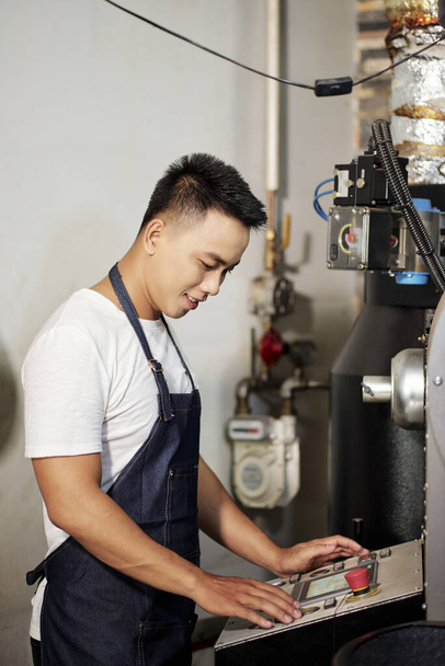 Улыбающийся вьетнамский молодой человек в джинсовом фартуке при эксплуатации новой кофеварки - Фото, изображение