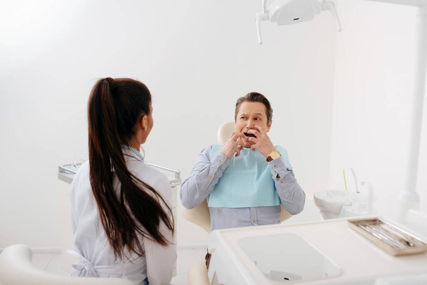 Rückansicht eines afrikanisch-amerikanischen Zahnarztes, der in der Nähe eines verängstigten Patienten sitzt und das Gesicht berührt  - Foto, Bild