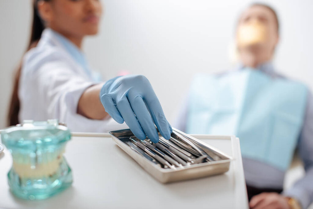 wybiórcze skupienie afrykańskiego amerykańskiego dentysty w rękawiczce lateksowej przyjmującego instrument stomatologiczny w pobliżu pacjenta  - Zdjęcie, obraz