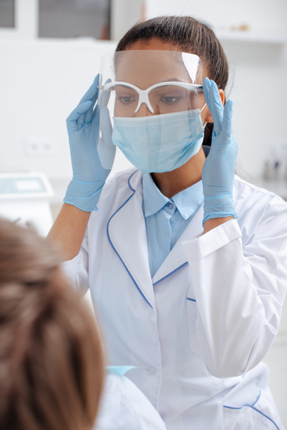 enfoque selectivo del dentista afroamericano en máscara médica tocando el protector facial cerca del paciente
  - Foto, Imagen