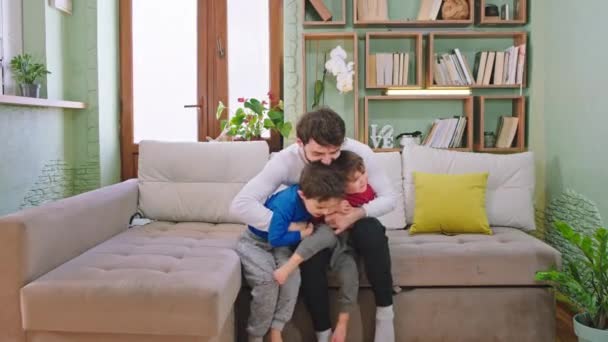 Νεαρός πατέρας και τα δύο παιδιά του με ένα μεγάλο χαμόγελο παίζοντας μαζί στον καναπέ στο σαλόνι απολαμβάνουν τη στιγμή μαζί μπροστά στην κάμερα - Πλάνα, βίντεο