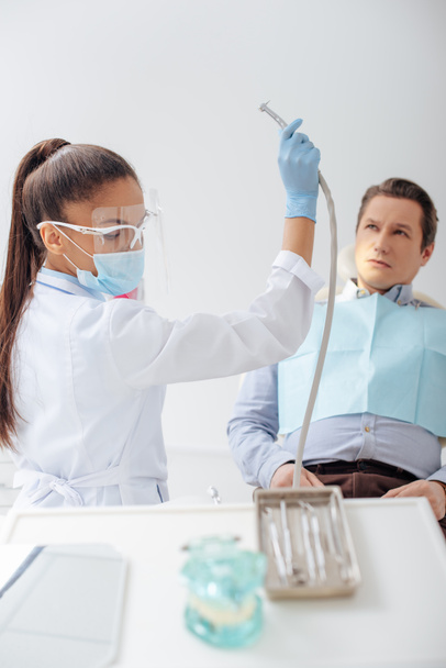 dentista afroamericano en máscara médica y escudo facial que sostiene el instrumento dental inoxidable cerca del hombre
  - Foto, Imagen