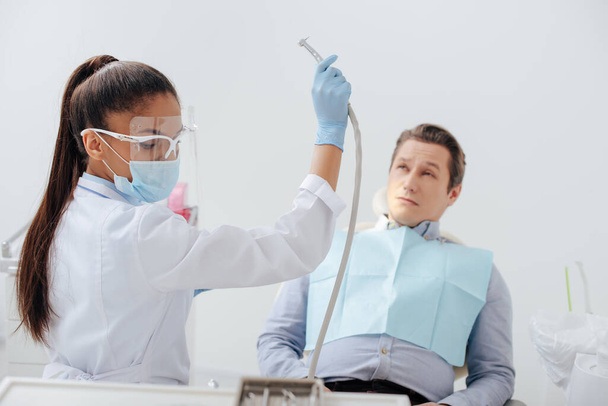 dentista afroamericano en máscara médica y escudo facial que sostiene el instrumento dental cerca del hombre
  - Foto, imagen