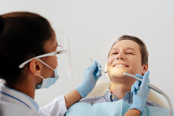 enfoque selectivo del dentista afroamericano en máscara médica y escudo facial que sostiene instrumentos dentales cerca de paciente alegre
  - Foto, Imagen