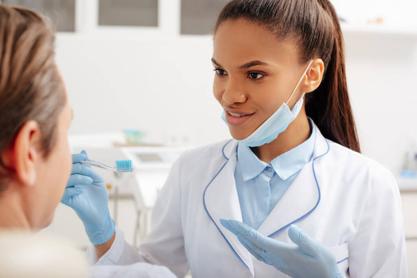 selective focus of happy african american dentist in latex gloves pointing with hand at toothbrush near patient  - Foto, Imagem