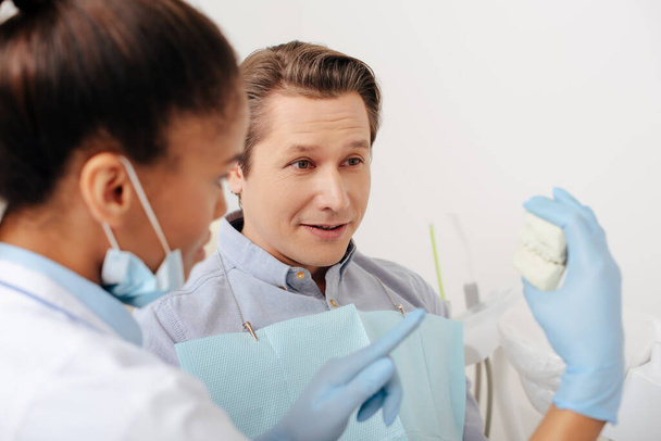 ラテックス手袋を着用したアフリカ系アメリカ人歯科医が患者の近くの歯モデルを指差して  - 写真・画像
