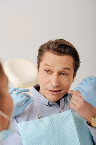 selective focus of african american dentist in latex gloves holding dental floss near handsome patient pointing with finger at face  - Foto, imagen