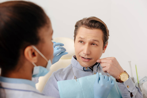 foco seletivo do dentista afro-americano em luvas de látex segurando fio dental perto do paciente apontando com o dedo no rosto
  - Foto, Imagem