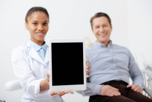 Neşeli Afrika kökenli Amerikalı dişçinin yanındaki mutlu hastanın seçici odağı elinde boş ekranlı dijital tablet tutuyor. - Fotoğraf, Görsel