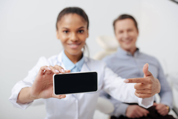 selective focus of cheerful african american dentist pointing with finger at smartphone with blank screen near happy patient  - Foto, Bild