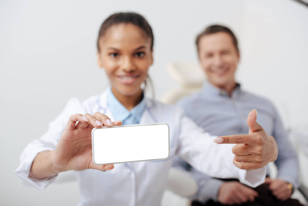 幸せな患者の近くに白い画面のスマートフォンで指を指している陽気なアフリカ系アメリカ人歯科医の選択的な焦点  - 写真・画像