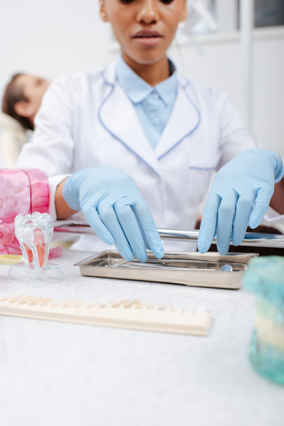 oříznutý pohled na afrického amerického zubaře v latexových rukavicích dotýkajících se nerezových zubních nástrojů poblíž pacienta  - Fotografie, Obrázek