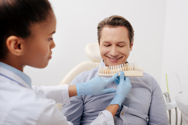 wybiórcze skupienie afrykańskiego amerykańskiego dentysty trzymającego paletę zębów w pobliżu szczęśliwego pacjenta w klinice  - Zdjęcie, obraz