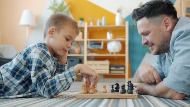 Smart child playing chess with dad at home lying on floor together having fun - Кадры, видео