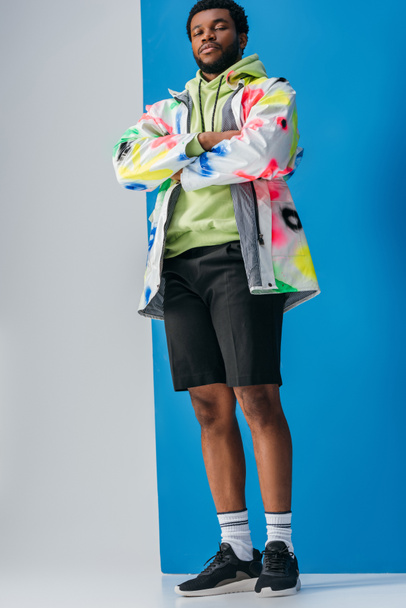 fashionable african american man posing with crossed arms in colorful futuristic look on grey and blue - Foto, Imagem