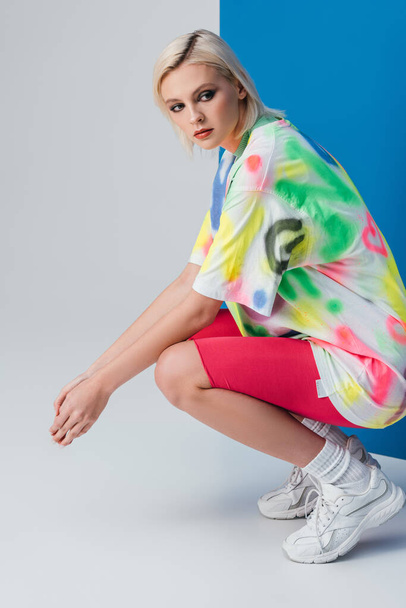 fashionable blonde woman posing in neon pink bike shorts and colorful t-shirt on grey and blue  - Foto, Imagen