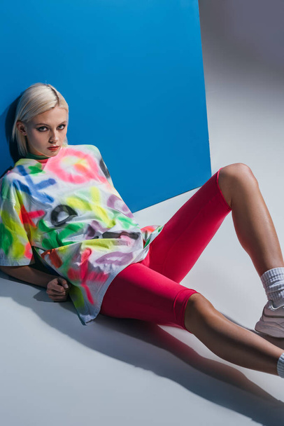 stylish blonde girl posing in neon pink bike shorts and colorful t-shirt on grey and blue  - Foto, immagini