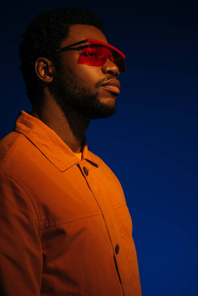 módní africký americký mužský model v futuristickém vzhledu a slunečních brýlích pózujících na modré v červeném světle  - Fotografie, Obrázek