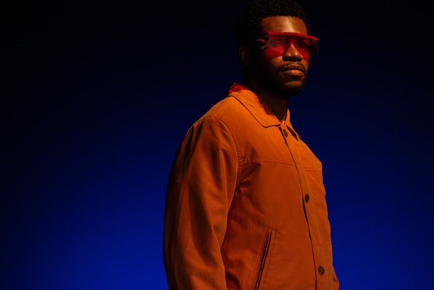 fashionable african american man in futuristic look and sunglasses posing on blue in red light  - Valokuva, kuva