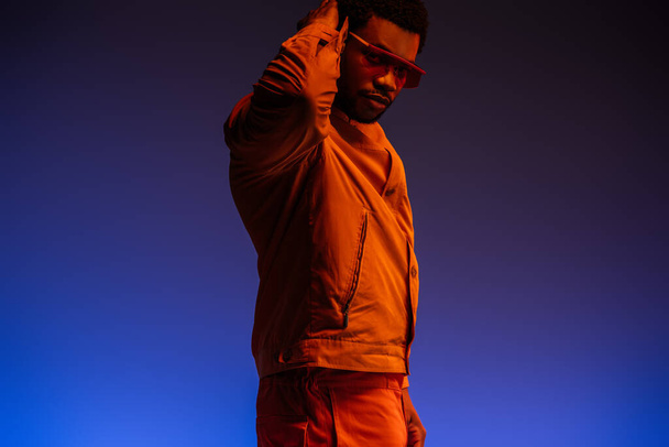 young african american man in futuristic look and sunglasses posing on blue in red light  - Foto, Imagen