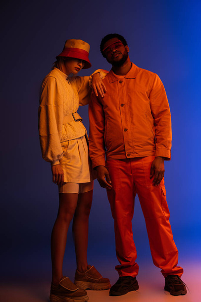 stylish interracial couple posing in futuristic look on blue in orange light - Фото, изображение