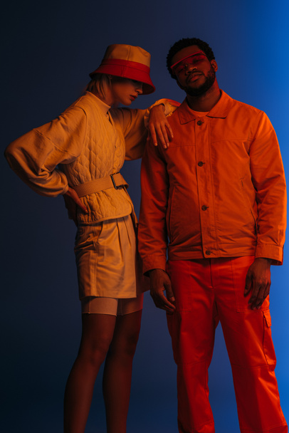 interracial couple in futuristic clothes, sunglasses and hat posing on blue in orange light - Foto, Imagen