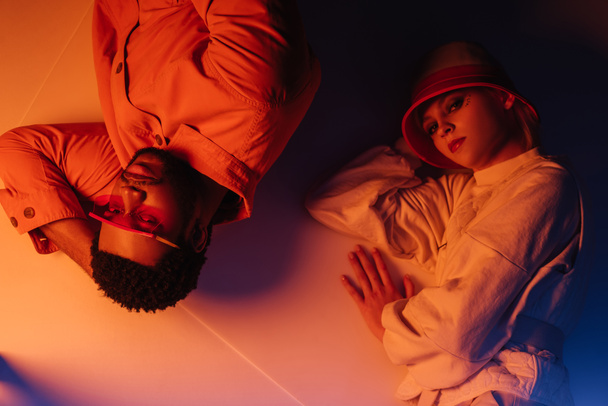 top view of interracial couple in futuristic clothes lying in orange light  - Foto, immagini
