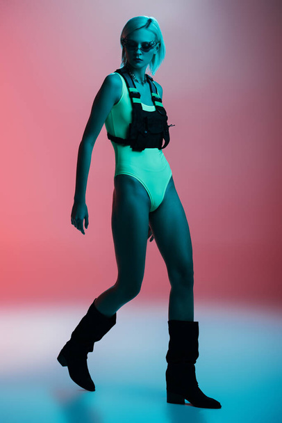 atractivo modelo de moda en traje de cuerpo futurista y gafas de sol en forma de fuego posando en rosa en luz azul
  - Foto, imagen