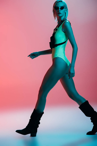 fashionable young model in futuristic leotard and fire-shaped sunglasses posing on pink in blue light  - Foto, Imagem