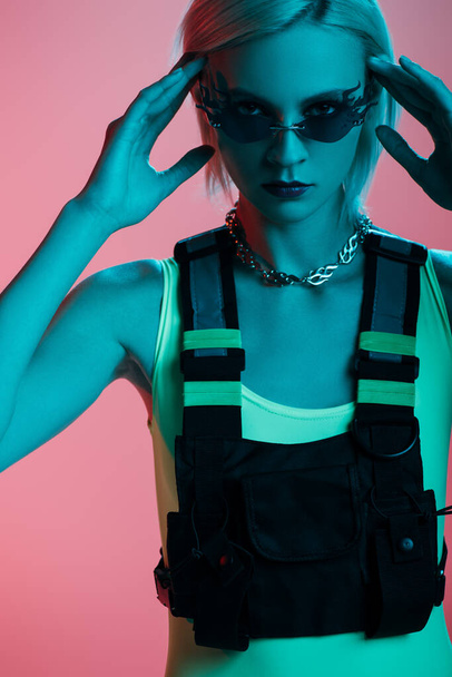 modelo joven de moda en traje de cuerpo futurista y gafas de sol en forma de fuego posando en rosa en luz azul
  - Foto, imagen