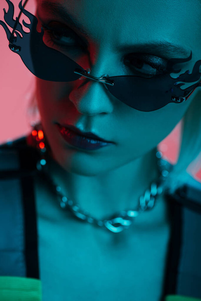 stylish female model in futuristic leotard and fire-shaped sunglasses posing isolated on pink in blue light  - Φωτογραφία, εικόνα