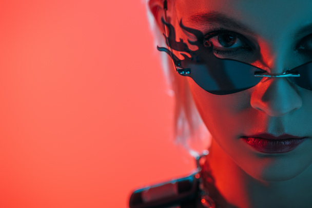 stylish futuristic girl posing in fire-shaped sunglasses on pink in red light - Фото, изображение