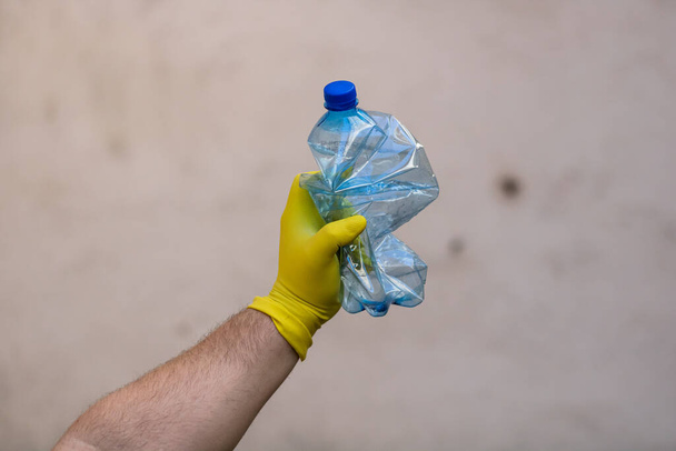 Mano con guante de goma que sostiene la botella de plástico vacía aplastada aislada sobre un fondo borroso. Concepto de reciclaje y voluntariado. Contaminación ambiental y problema ecológico mundial
 - Foto, Imagen