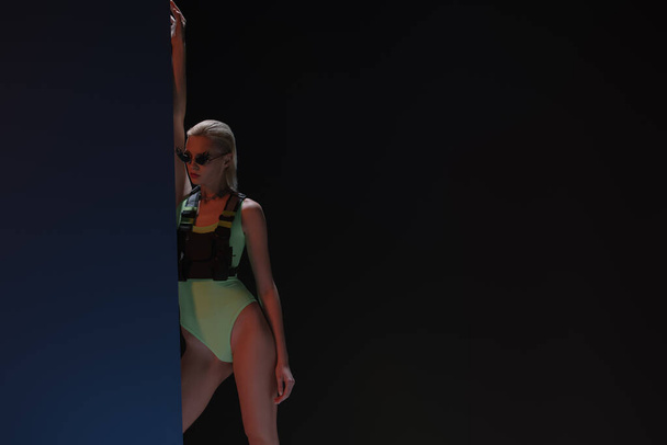 modisches Mädchen in futuristischem Body und feuerförmiger Sonnenbrille posiert auf dunklem  - Foto, Bild