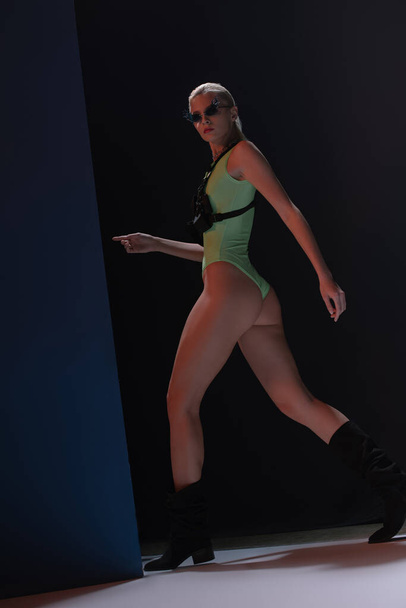 attractive fashionable girl in futuristic leotard and fire-shaped sunglasses posing on dark  - Foto, afbeelding