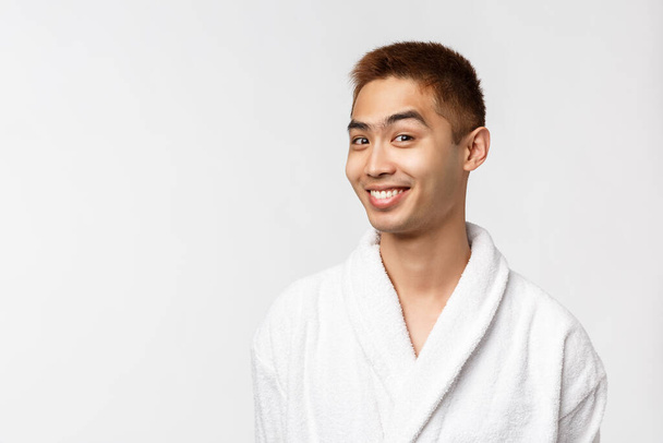 Концепция красоты, spa и отдыха. Дерзкий любопытный красивый азиатский мужчина в халате смотреть камера удивлена и заинтригована, улыбаясь, голова интересное предложение, в халате, белый фон
 - Фото, изображение