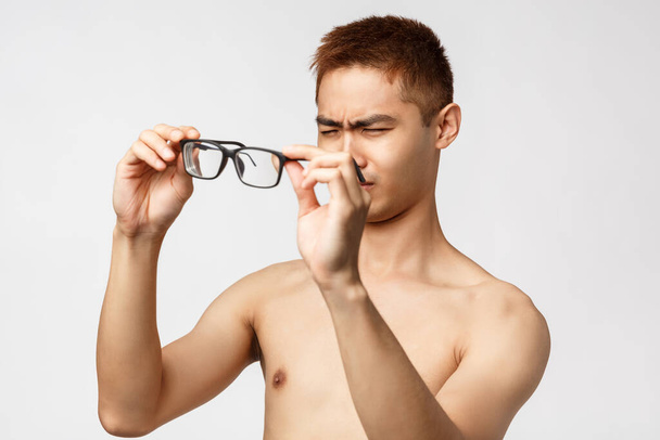 Beauty, people and lifestyle concept. Portrait of young handsome asian man with naked torso, waking up morning cant see anything without glasses, squinting put on eyewear, white background - Foto, Bild