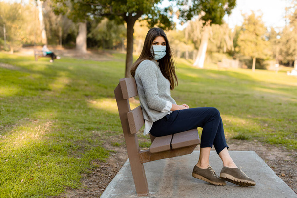 Mujer joven al aire libre con mascarilla médica, distanciamiento social, sentada en un banco, aislada de otras personas
. - Foto, imagen