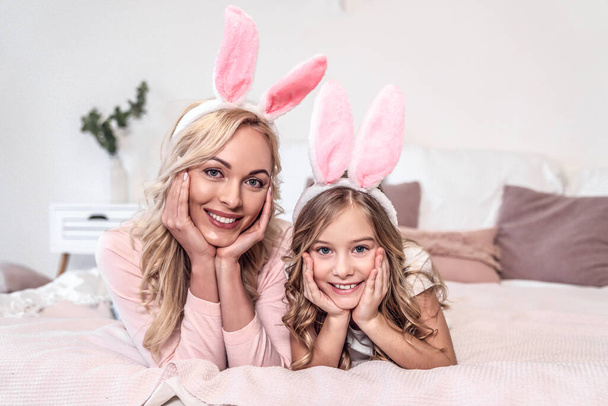 Atraktivní mladá žena se svou roztomilou dceruškou ležící spolu na posteli v králičích uších. Oslavy Velikonoc - Fotografie, Obrázek