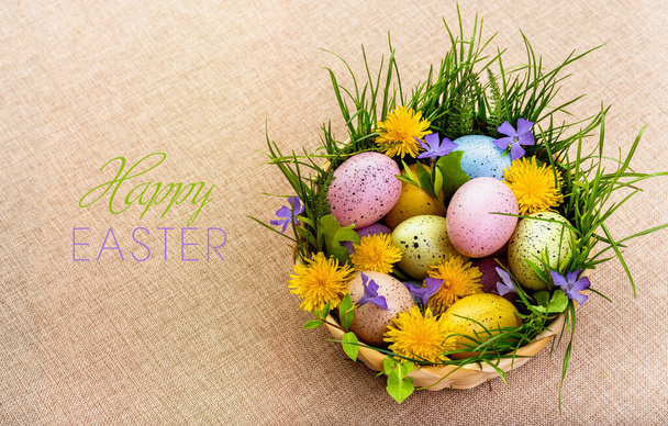 イースターだ。イースターの春の休日におめでとうございます。キャンバスを背景に春の花や草で飾られた籠の中の多色の卵. - 写真・画像