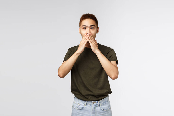 Surprised and amused asian man gasping, cover mouth and look camera impressed, express astonishment and happiness for friend, hear great news, standing grey background pleased - Photo, Image