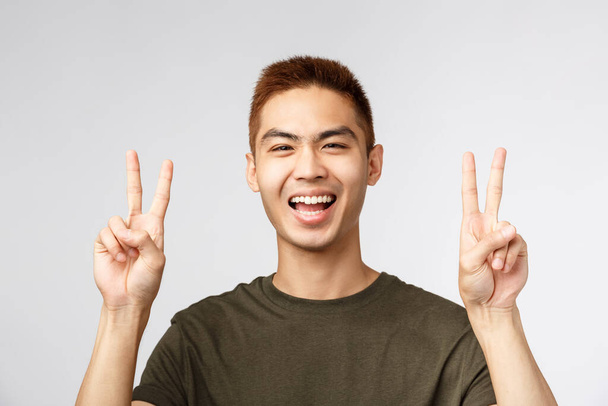 Lidé, různé výrazy a životní styl. Detailní záběr portrét radostného pohledného mladého studenta, japonský kluk se cítí dobře, připravuje se na dovolenou, ukazuje mírové znamení a usměje se - Fotografie, Obrázek