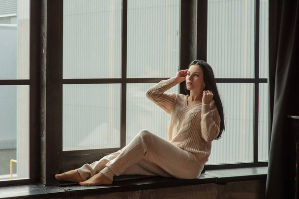 Young attractive woman posing in fashionable clothes near the window. Girl at the window in a beige knitted suit. Brunette, beautiful young girl, portrait. - Photo, Image