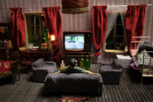 A realistic dollhouse living room with furniture and window at night. Artwork table decoration with handmade realistic dollhouse. Man wathing retro style Television in dark room. Selective focus. - Foto, imagen