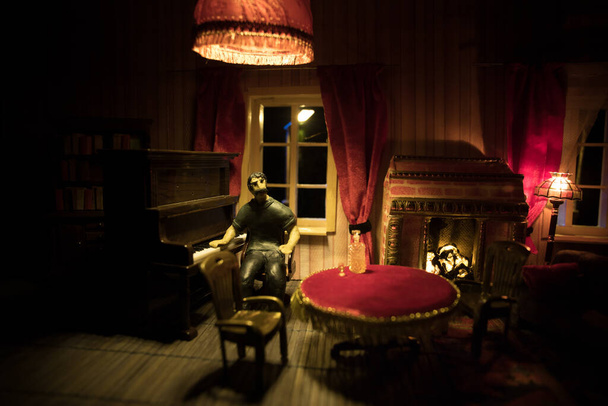 Una casa de muñecas realista sala de estar con muebles y ventana por la noche. Hombre sentado al piano viejo en cuarto oscuro. Enfoque selectivo
. - Foto, imagen