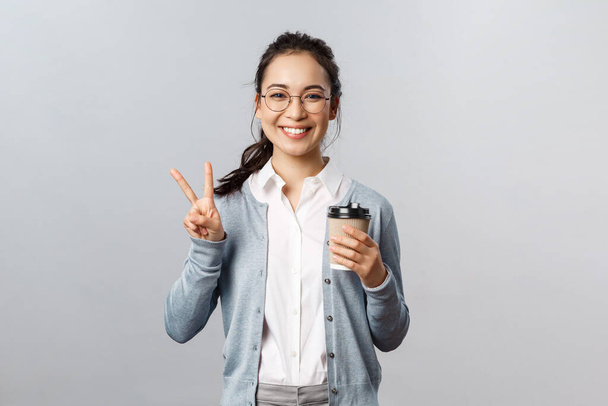 Офисный образ жизни, бизнес и концепция людей. Оптимистичная, дружелюбно выглядящая азиатка показывает знак мира, улыбается Кавайи, наслаждаясь утренней свежей чашкой кофе из местного кафе, стоять на сером фоне
 - Фото, изображение