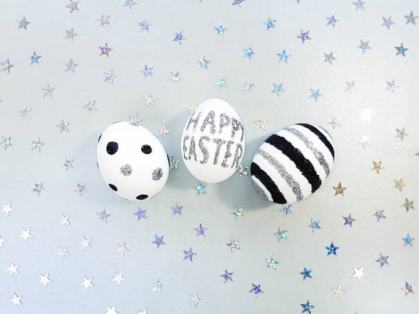 Composition of silver glitter inscription Happy Easter on a white egg and metallic stars spangles sequins. Eggs with black and silver lines and polka dots. Flat lay, top view, close up, copy space - Photo, Image