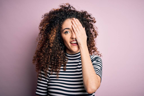 Молодая красивая женщина с вьющимися волосами и пирсинг носить случайные полосатые футболки покрывая один глаз рукой, уверенная улыбка на лице и удивление эмоций
. - Фото, изображение