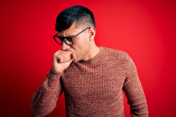 Jonge knappe Latijns-Amerikaanse man draagt een nerd bril over rode achtergrond die zich onwel voelt en hoestend als symptoom voor verkoudheid of bronchitis. Gezondheidszorgconcept. - Foto, afbeelding
